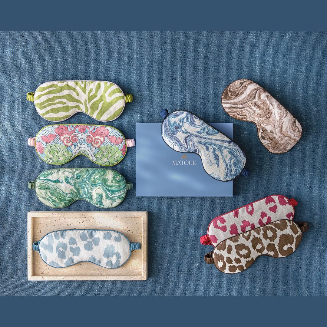 Six patterned sleep masks with blackout lining surround a blue Matouk Schumacher box on a blue fabric background.