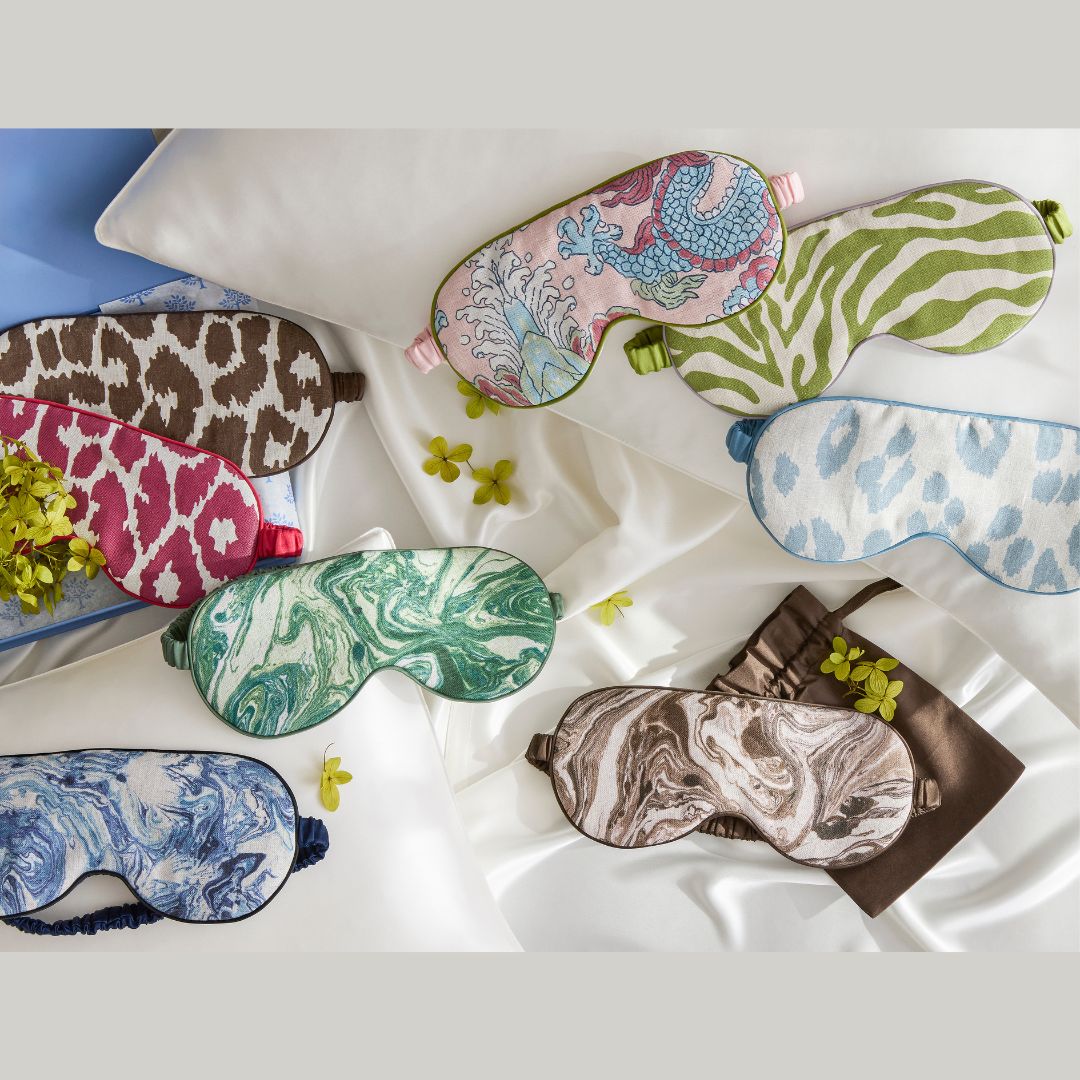 Several patterned sleep masks with blackout lining by Matouk Schumacher are arranged on a bed with white sheets and brown pillowcases.