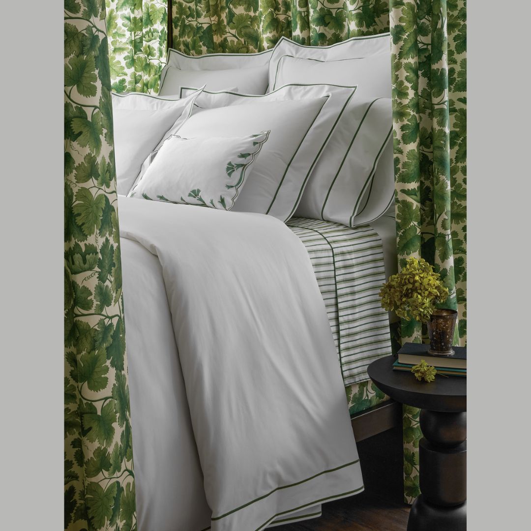 Neatly made bed with white bedding and green accents against a green leafy curtain.