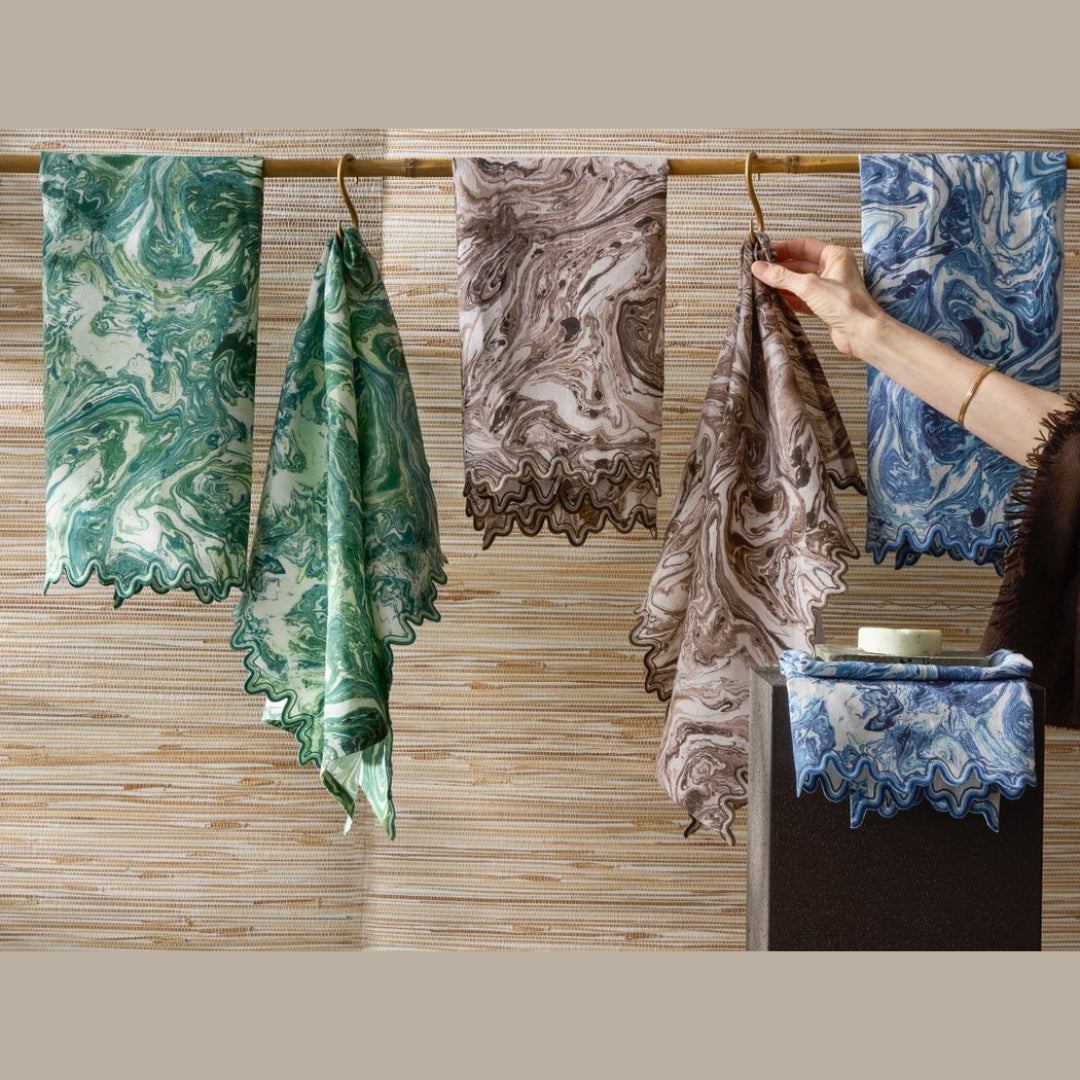 Five marbled-patterned scarves hang on a rod; reminiscent of marbled linen guest towels, a hand holds one brown scarf.