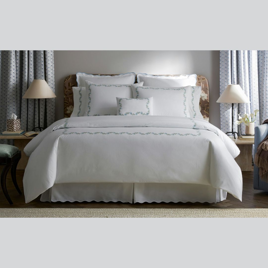 A neatly made bed with 520 thread count Ceylon percale and embroidered floral vine, flanked by lamps and patterned curtains.