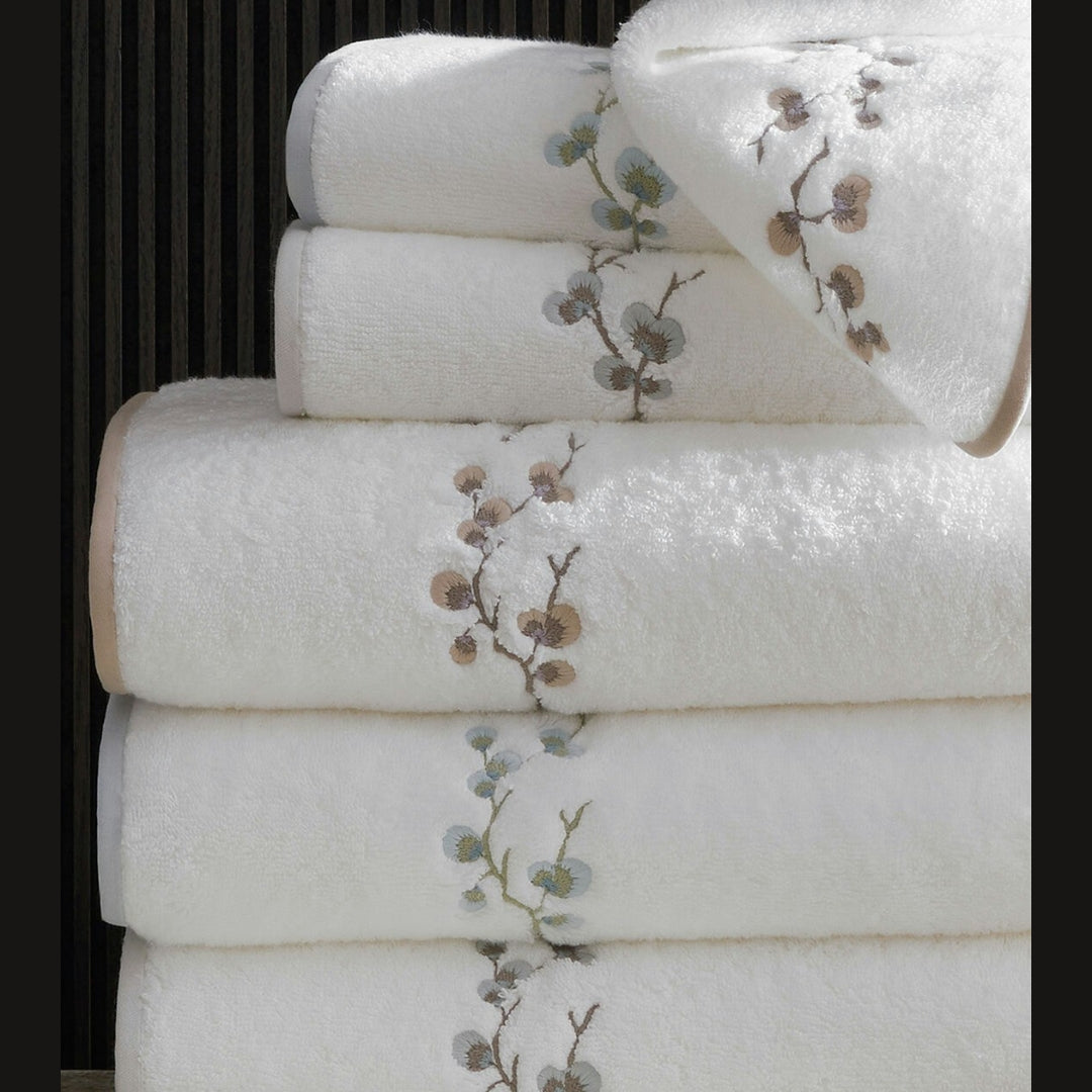 A stack of white plush towels with delicate floral embroidery in muted brown and blue-gray tones, photographed against a dark vertical slatted background.