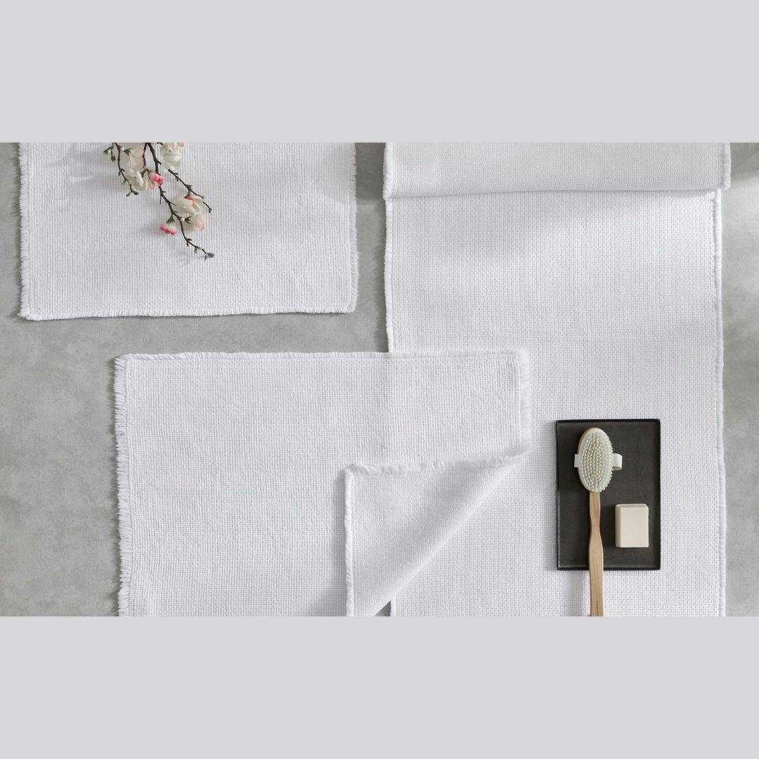 White textured towels with frayed edges rest beside a lightweight woven cotton bath rug and soap on a gray surface.