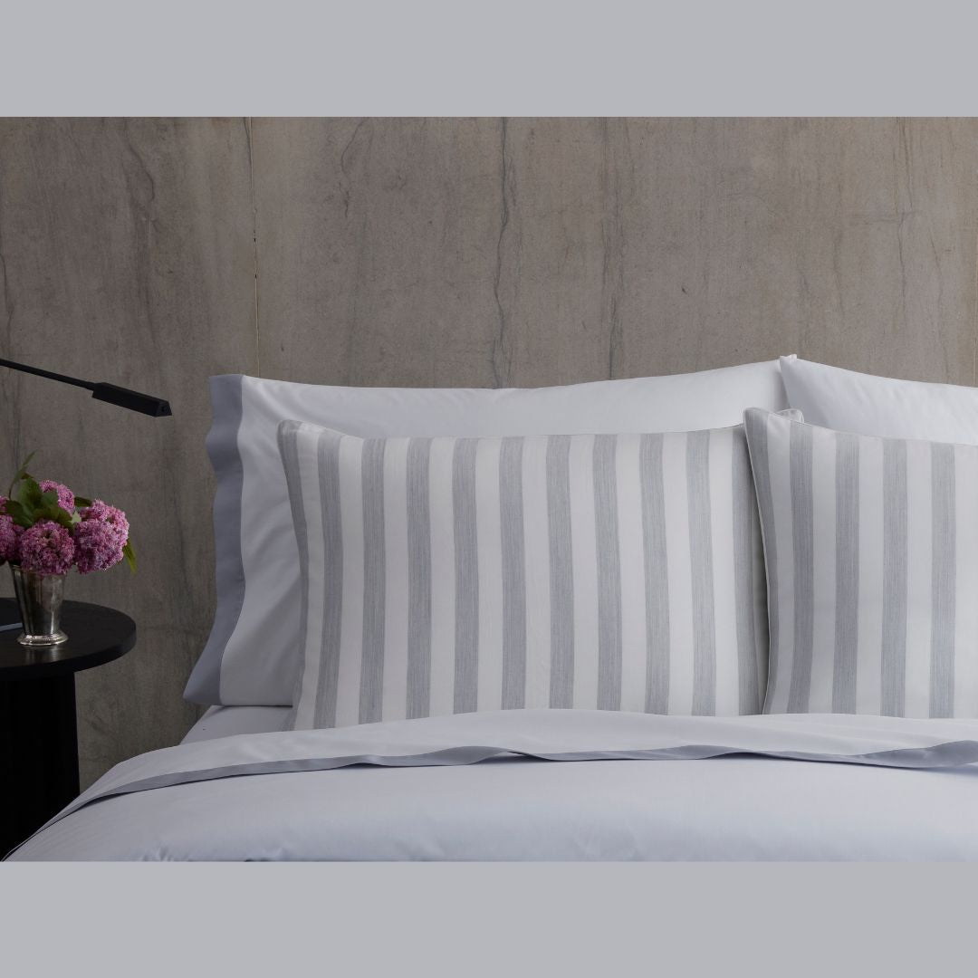Bed with white and gray striped pillows, white sheets, luxury duvet covers, a dark nightstand, and pink flowers in a vase.