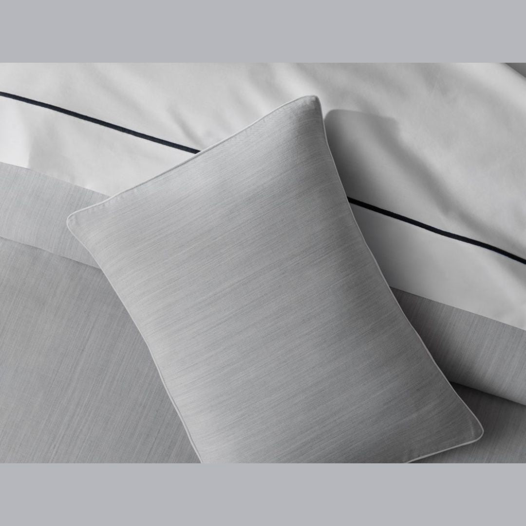A light gray pillow rests on Matouk Lana Bedding, with a white sheet featuring a thin dark line above it.
