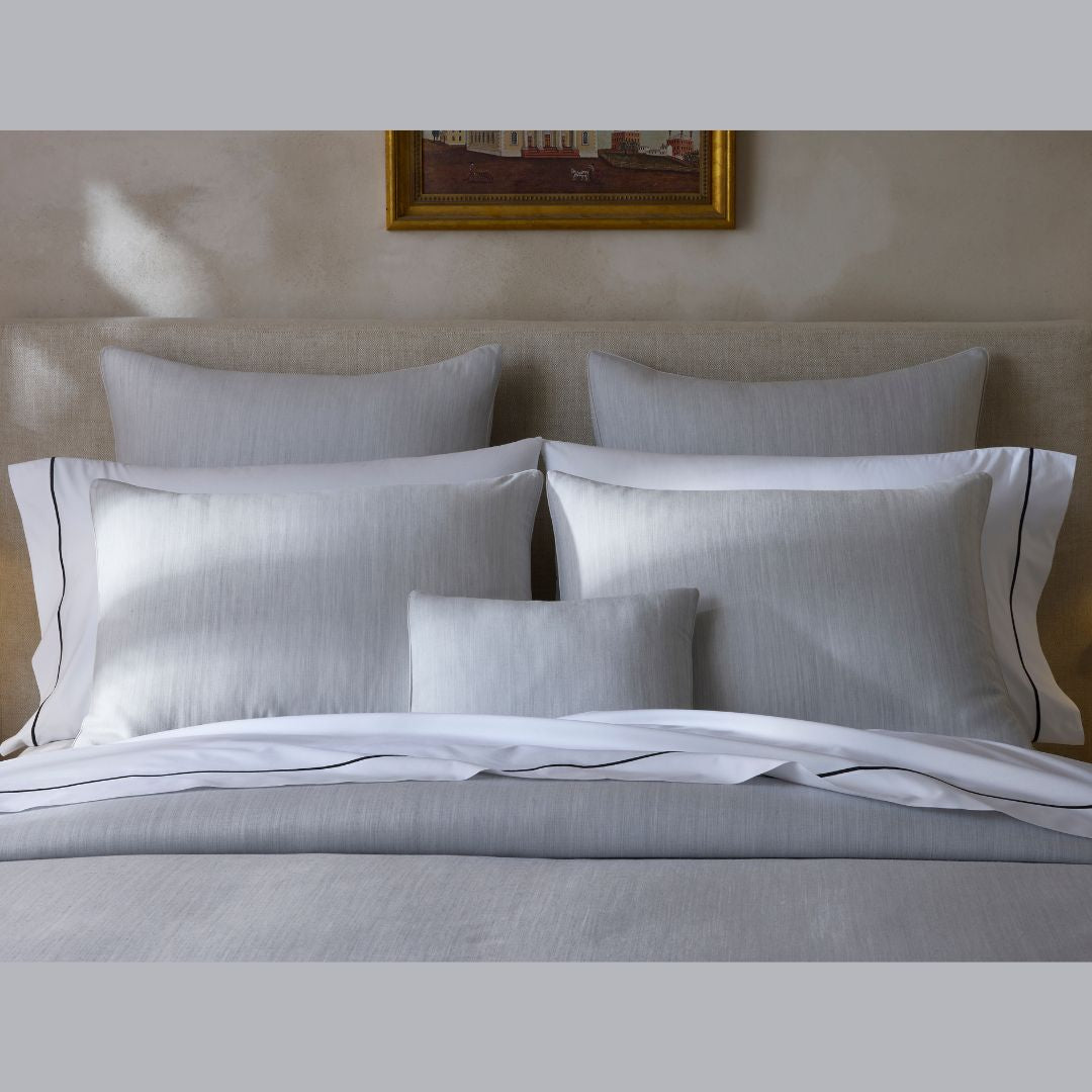 A neatly made bed features Matouk Lana Bedding, gray and white pillows, white sheets, and a beige upholstered headboard.