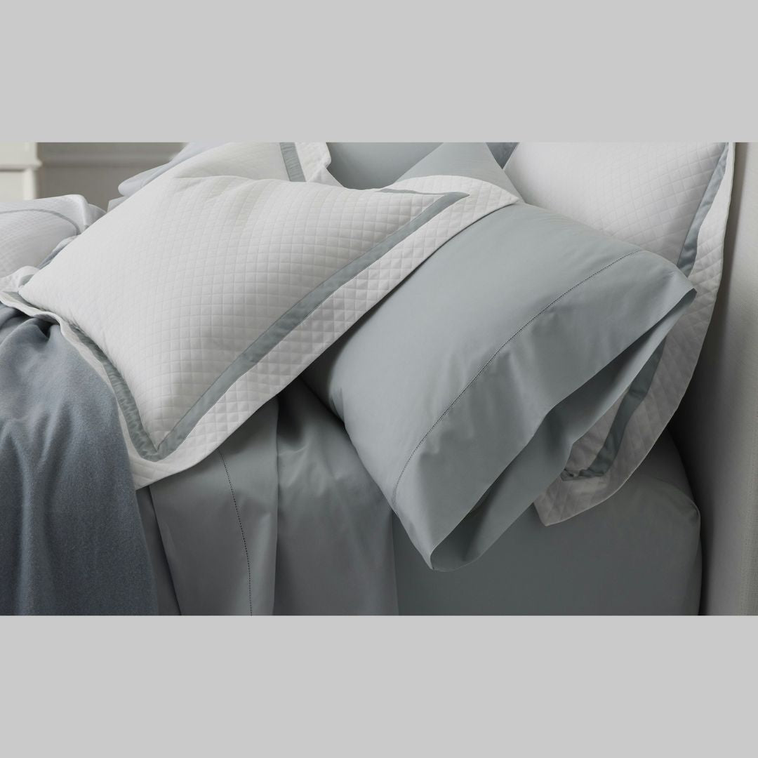 Light blue bedding with two white pillows featuring grey trim, crafted from Matouk Sheeting for luxe comfort.