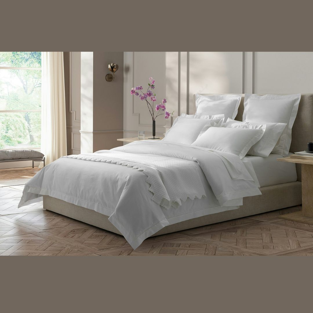 A neatly made bed with Milano 600 Thread Count white bedding, orchid on the nightstand, and sunlight streaming in.