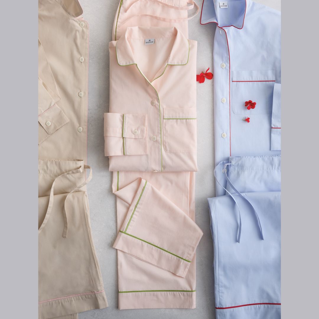 Three pairs of pajama sets arranged on a light gray background: a beige short-sleeve top and shorts set on the left, a pale pink long-sleeve pajama set with green piping in the center, and a light blue long-sleeve pajama set with red piping on the right. Small red flower props are placed near the center and right sets.