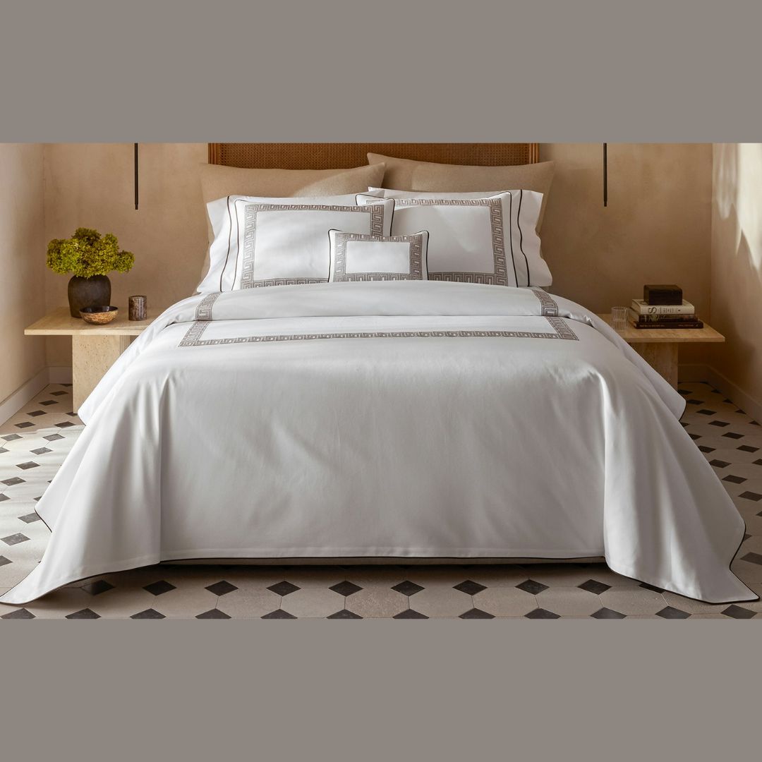 A neatly made bed with a luxury cotton coverlet featuring Greek key bedding, flanked by nightstands in a neutral room.