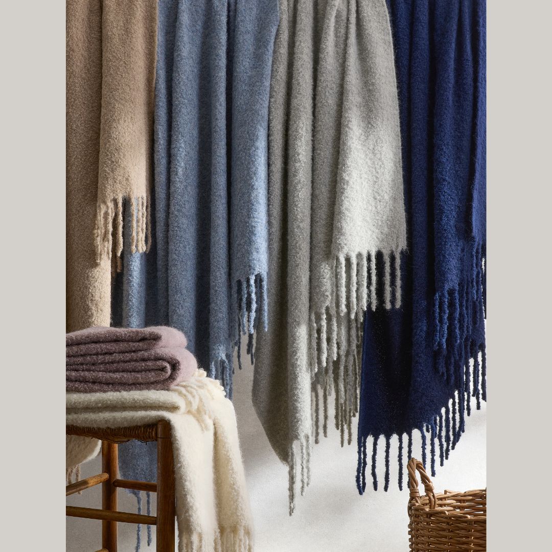 Cozy alpaca throws in beige, blue, gray, and navy hang with fringed edges; folded blankets sit on a wicker bench.