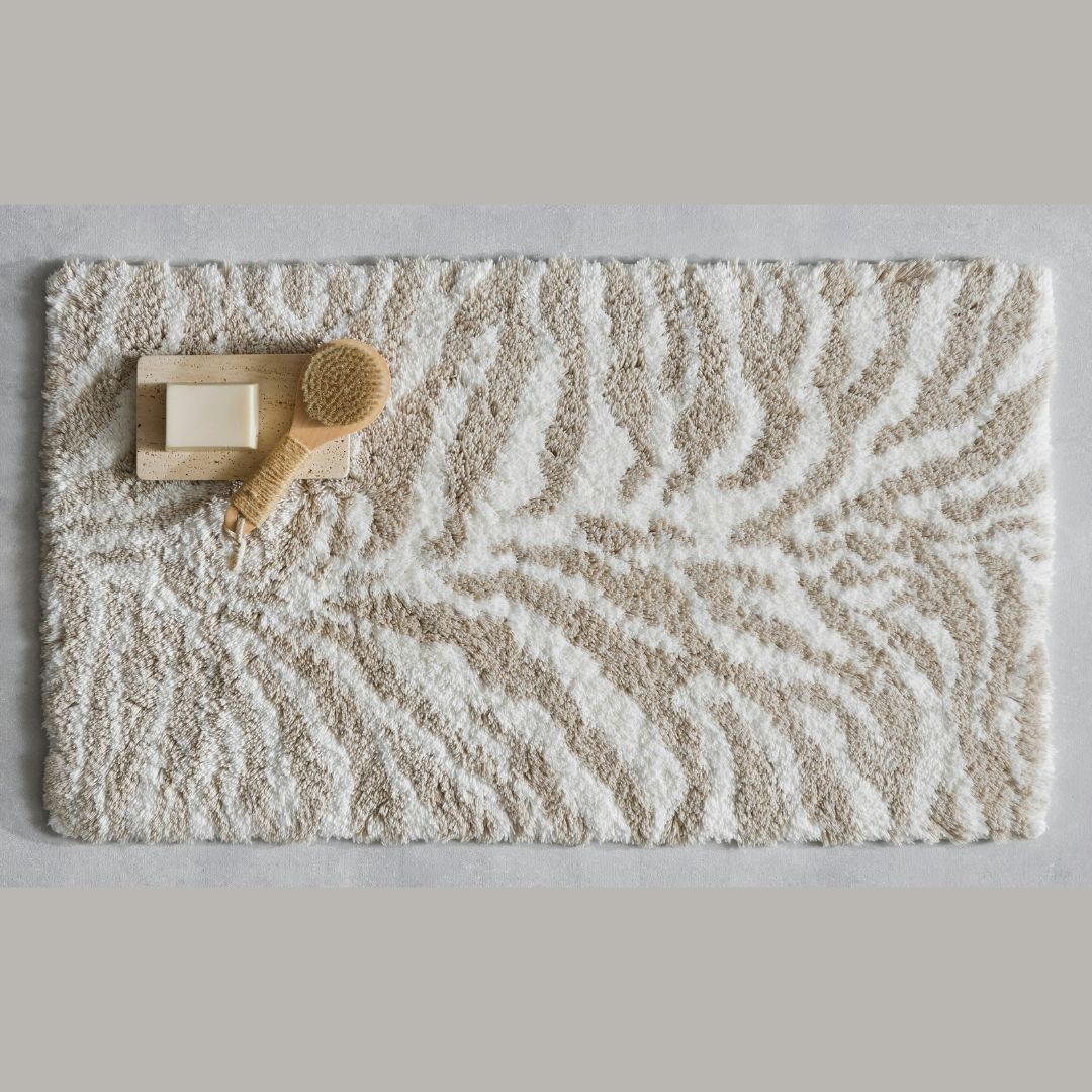 Beige and white Matouk Regine Rug bath mat with zebra pattern, wooden soap dish, bar of soap, and wooden brush.