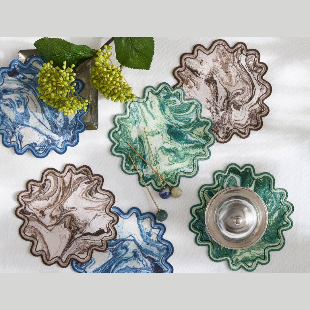 Six scalloped ceramic coasters with marbled patterns in blue, green, and brown, arranged on a white textured background with decorative grapevine props and a small silver bowl.