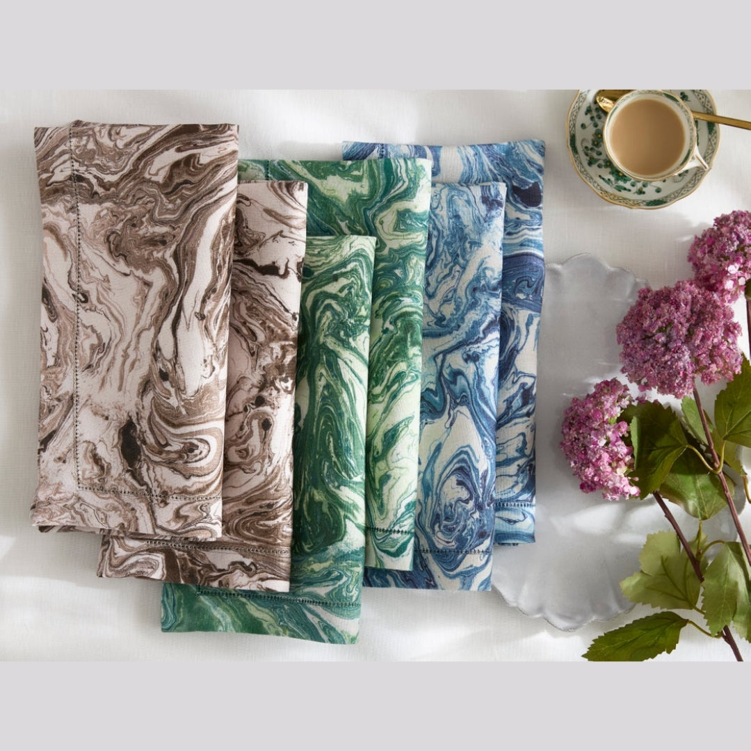 Six folded fabric napkins with marble patterns in brown, green, and blue are arranged on a white surface next to a cup of coffee and pink flowers.