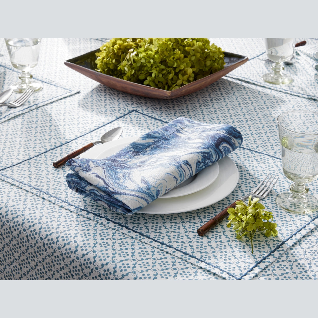 A styled table setting featuring a folded blue and white marbled napkin on a white plate, set on a blue and white patterned tablecloth. A matching patterned placemat is visible, along with a glass, silverware, and a decorative bowl of green hydrangeas in the background.
