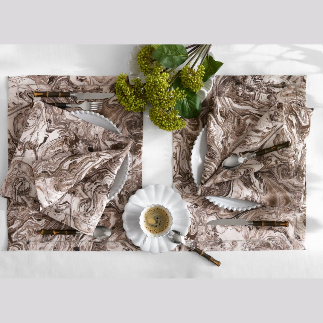 Two matching marble-patterned placemats styled on a white table with white plates, silver cutlery, a small cup of coffee, and green grapevine stems.