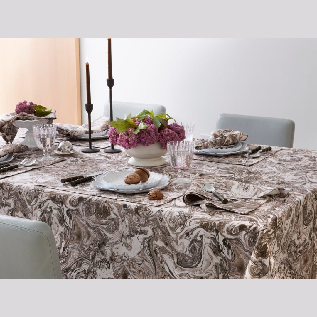 A patterned tablecloth with a brown and beige marble-like swirl design draped over a dining table, styled with plates, cutlery, candles, and a floral centerpiece.