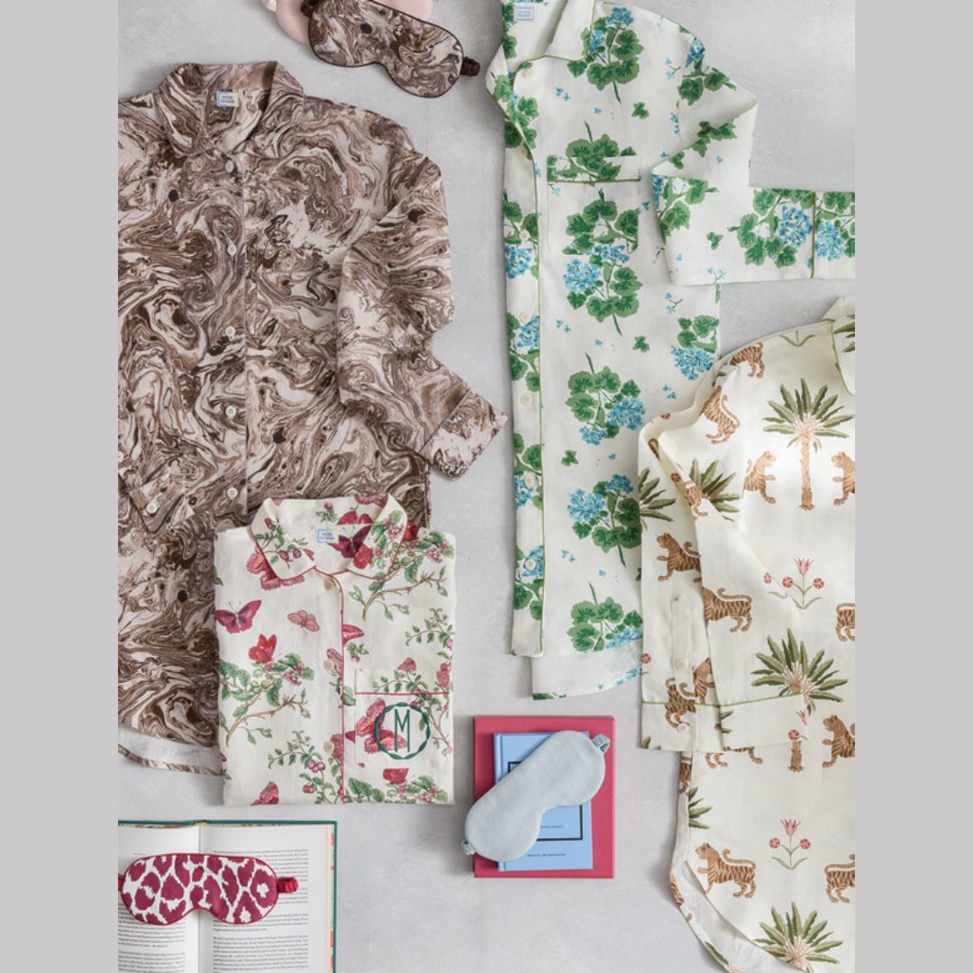 A styled flat lay of multiple pajama sets and matching accessories on a light gray background. The collection includes a brown marbled pajama top, a cream pajama set with green and blue floral print, a cream pajama set with pink birds and green foliage, a cream pajama set with brown animal and palm tree print, a matching sleep mask, and a small book with a giraffe-print eye mask on it.