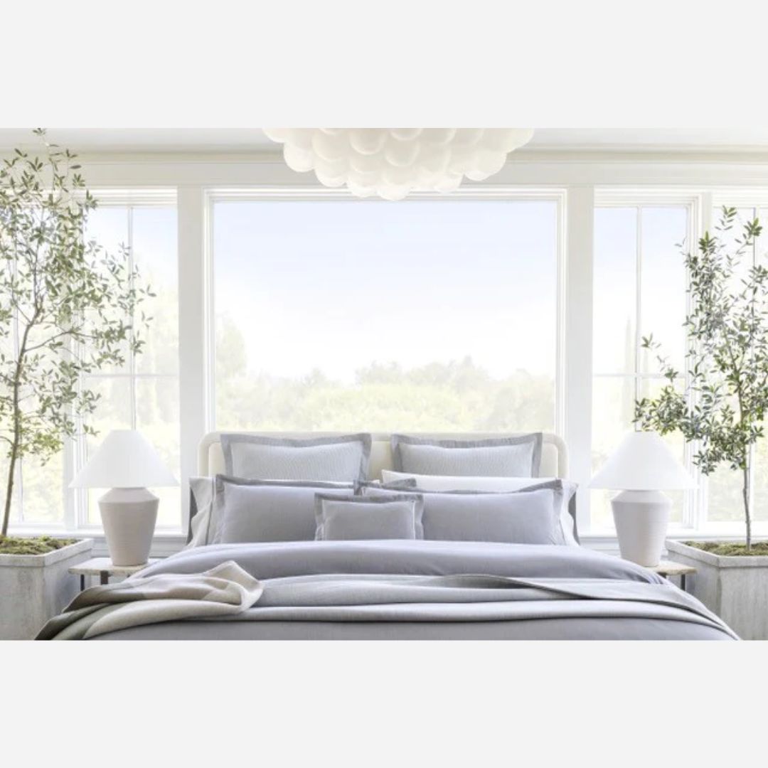 A neatly made bed with gray Italian woven bedding, two white lamps, and potted trees in front of large windows.
