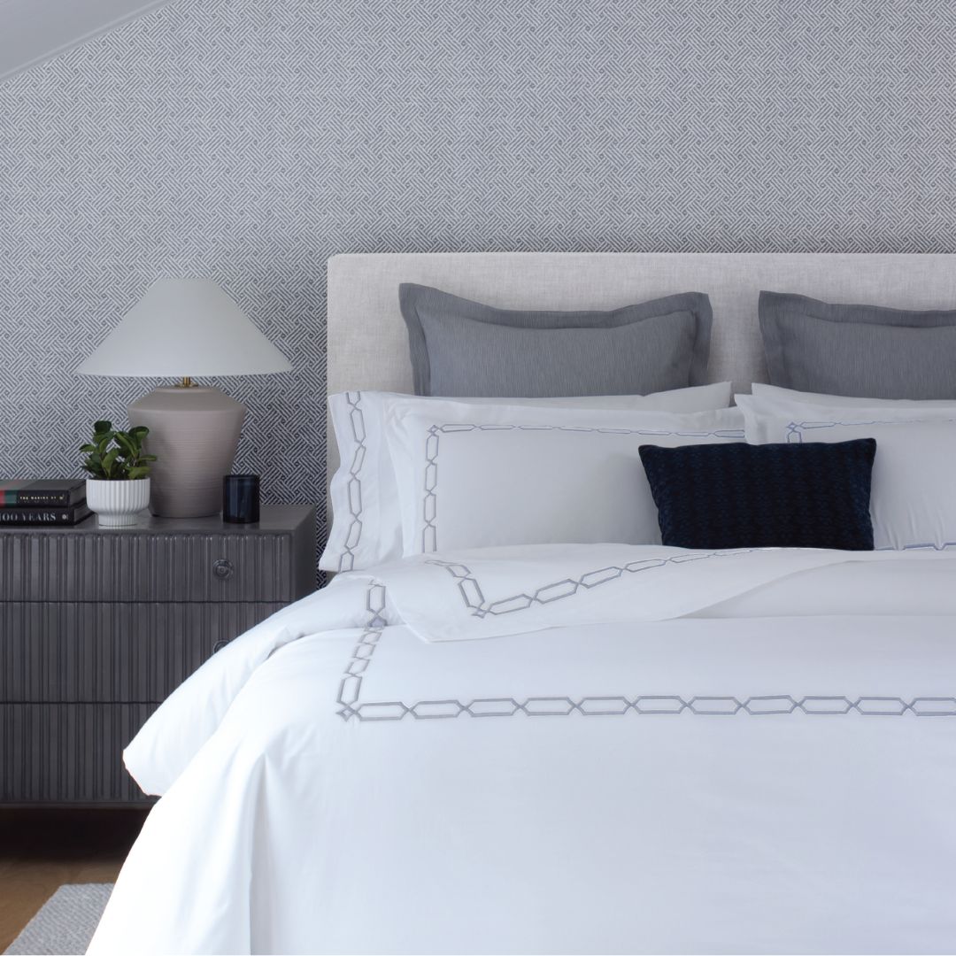 A neatly made bed with luxury bedding, gray pillows, a navy cushion, and a bedside table with a lamp and a plant.