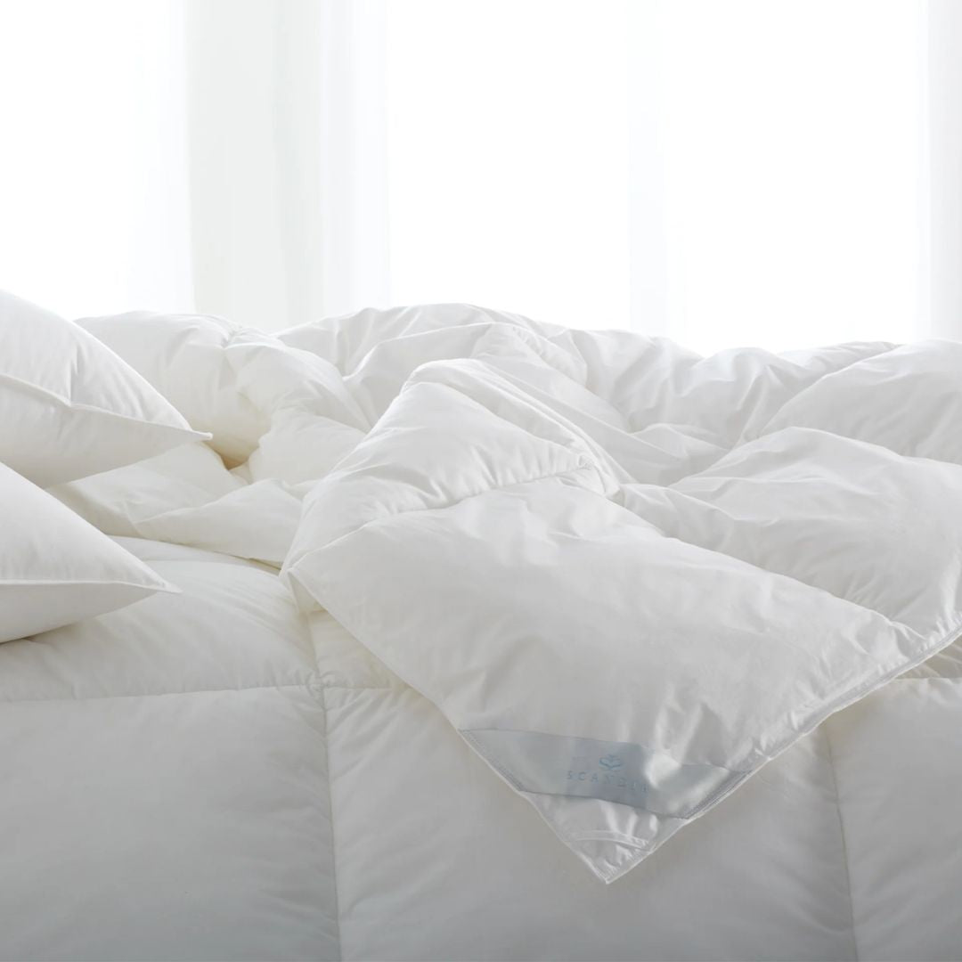 A white, fluffy synthetic fill comforter and pillows are arranged on a bed in a bright room with sheer curtains.