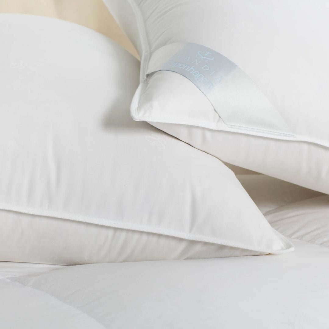 Two European white down pillows with satin trim are stacked on a white quilted surface, featuring a fabric brand tag.