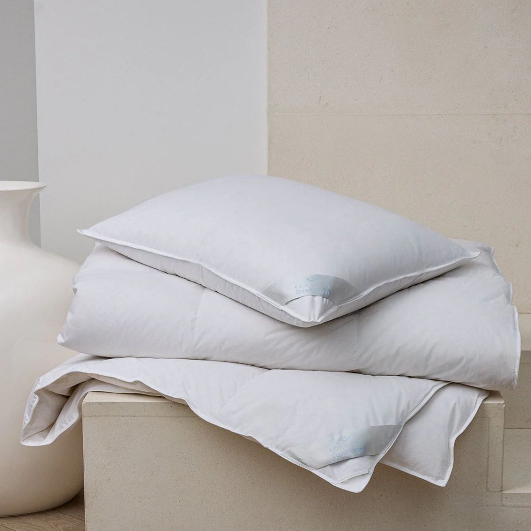 A white pillow and Scandia Home comforter with cotton cambric ticking are neatly stacked next to a large white vase.