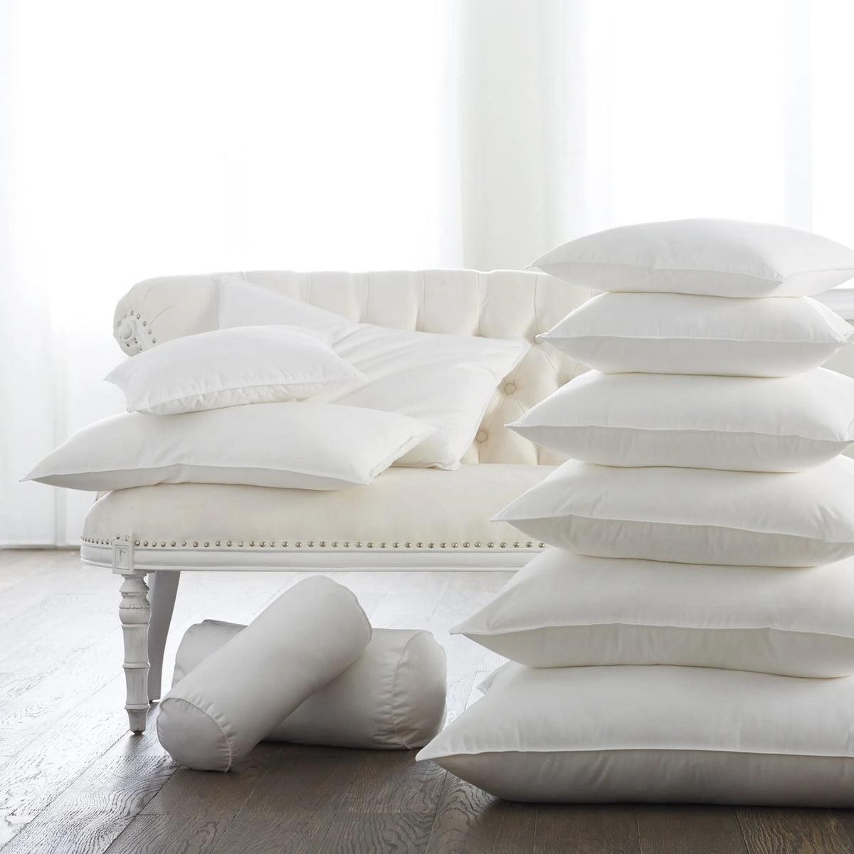 A white couch with feather-down blend pillows and two bolster pillows beside a tall stack of white pillows on the floor.