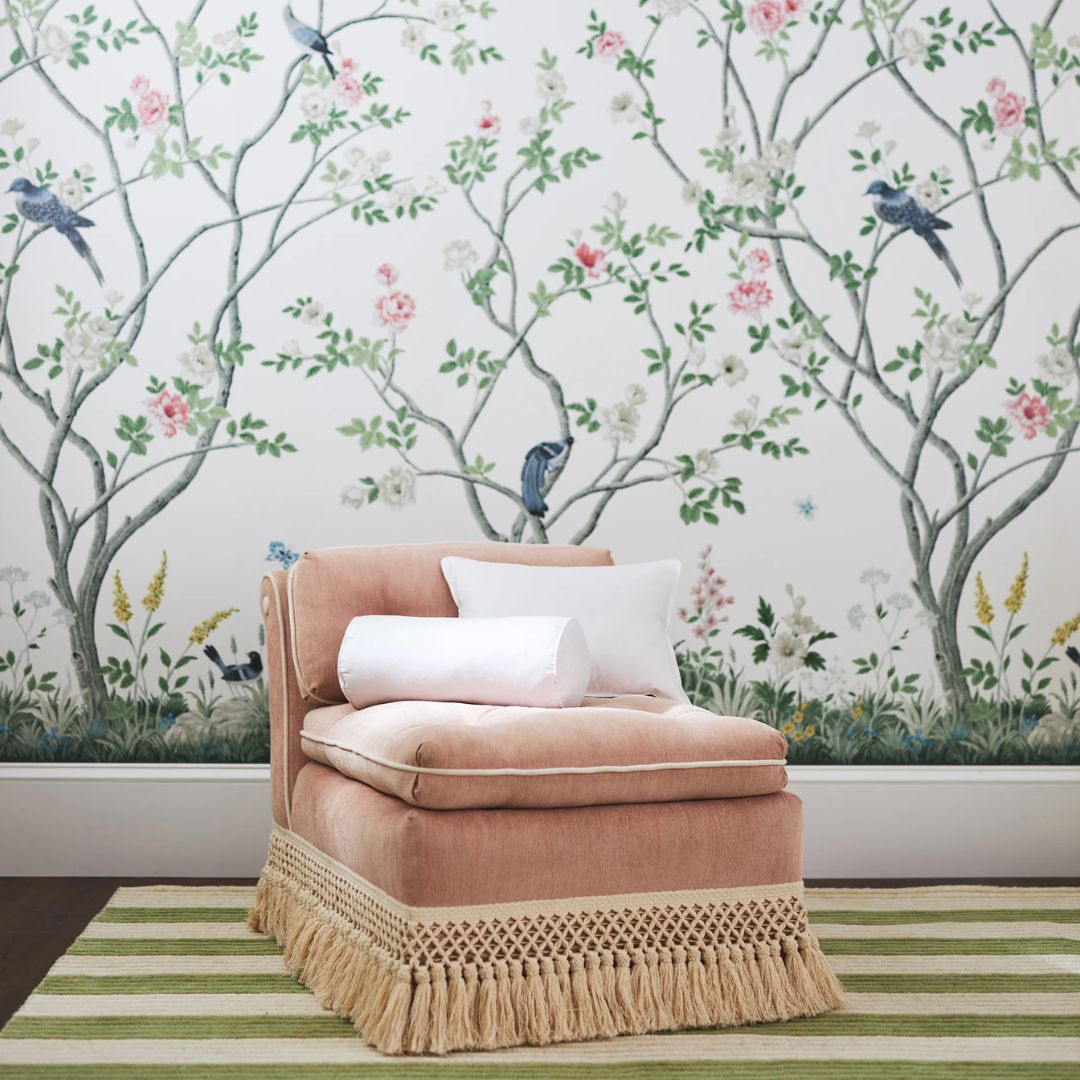 A pink chaise lounge with a decorative insert pillow sits on a striped rug, with floral and bird-patterned wallpaper behind it.