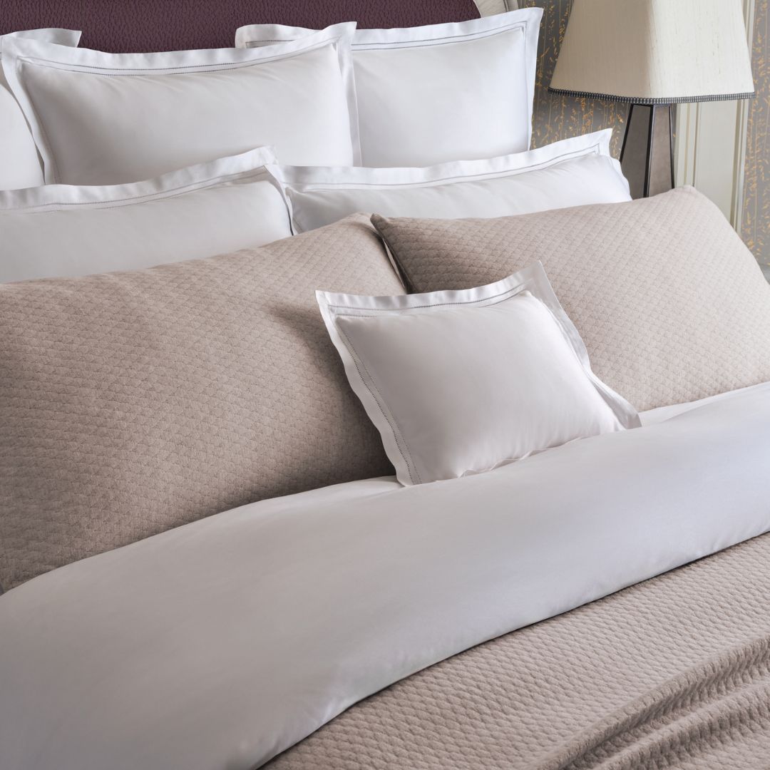 A neatly made bed with a luxury coverlet, white pillows, and beige quilted bedding in a softly lit bedroom.