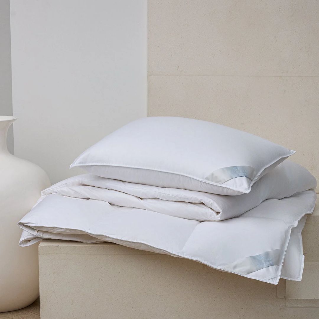 A Scandia Home pillow and folded white comforter rest on a beige surface beside a large white vase.