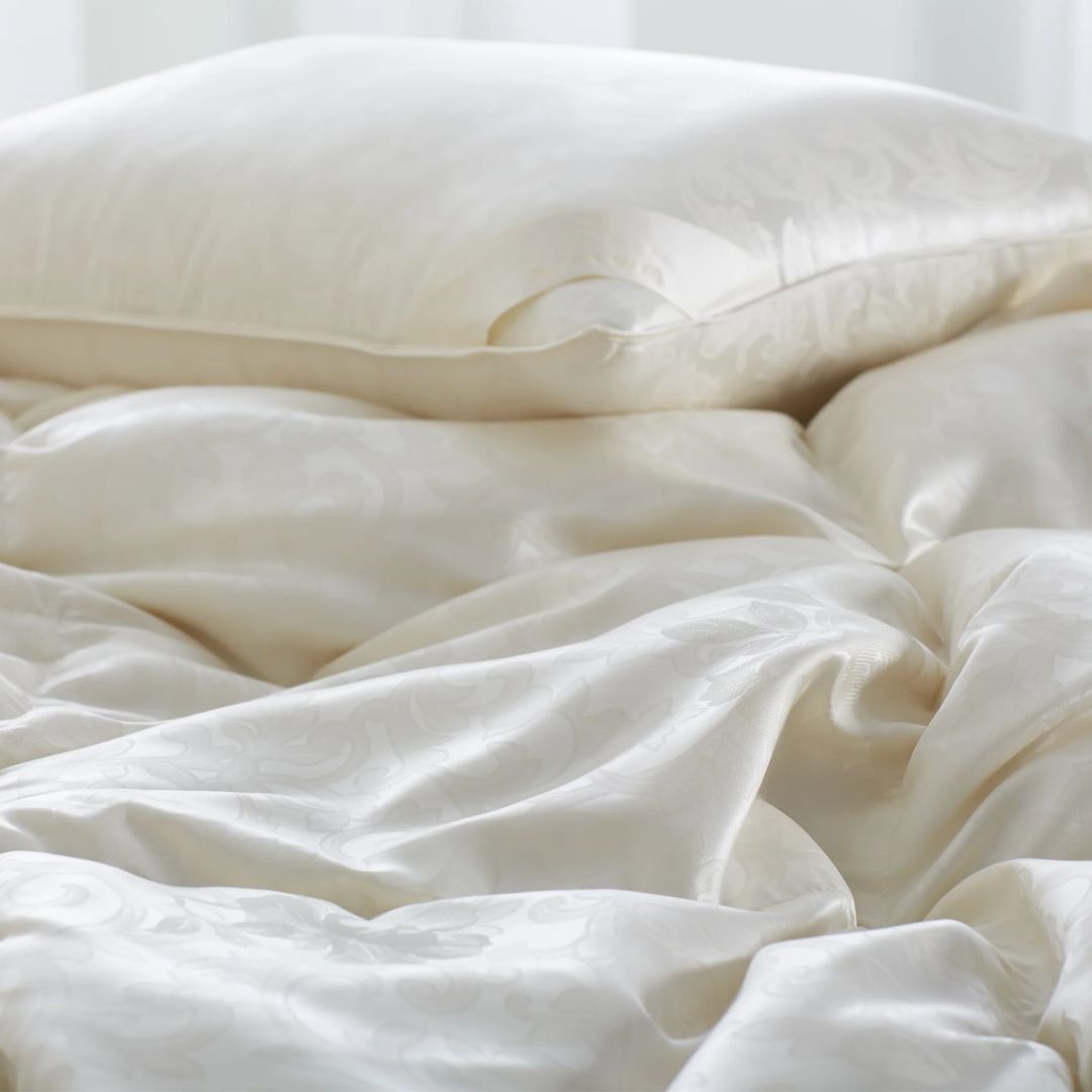 A close-up of a cream-colored silk damask pillow and luxury down comforter with a subtle floral pattern on a bed.