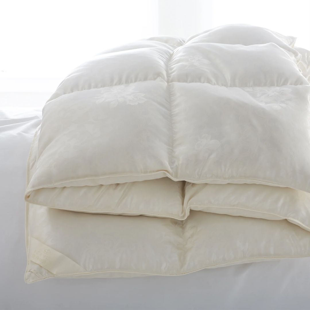 A folded luxury down comforter with a subtle floral pattern rests on a white bed.