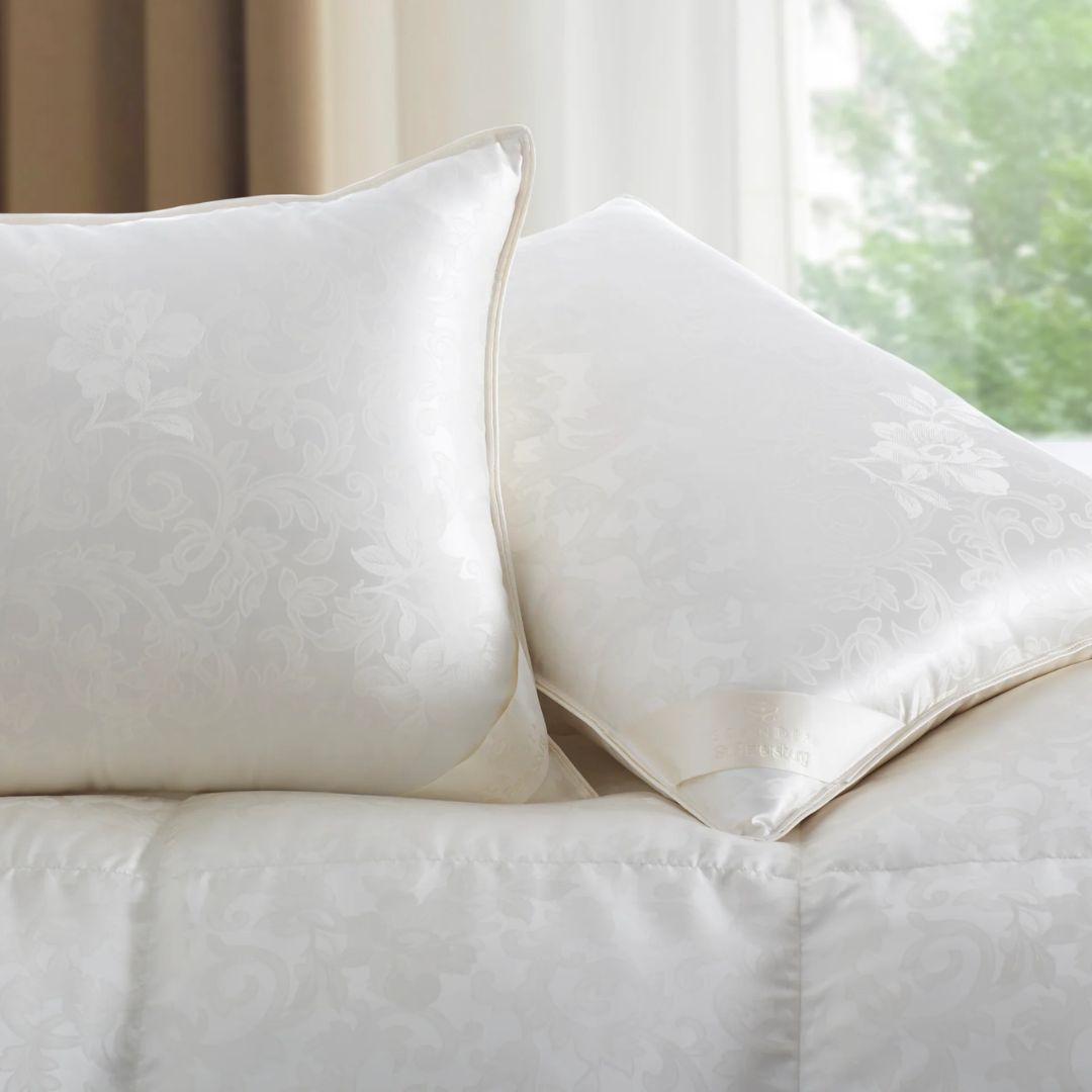 Two white luxury down pillows with a subtle floral pattern rest on a matching white bedspread.