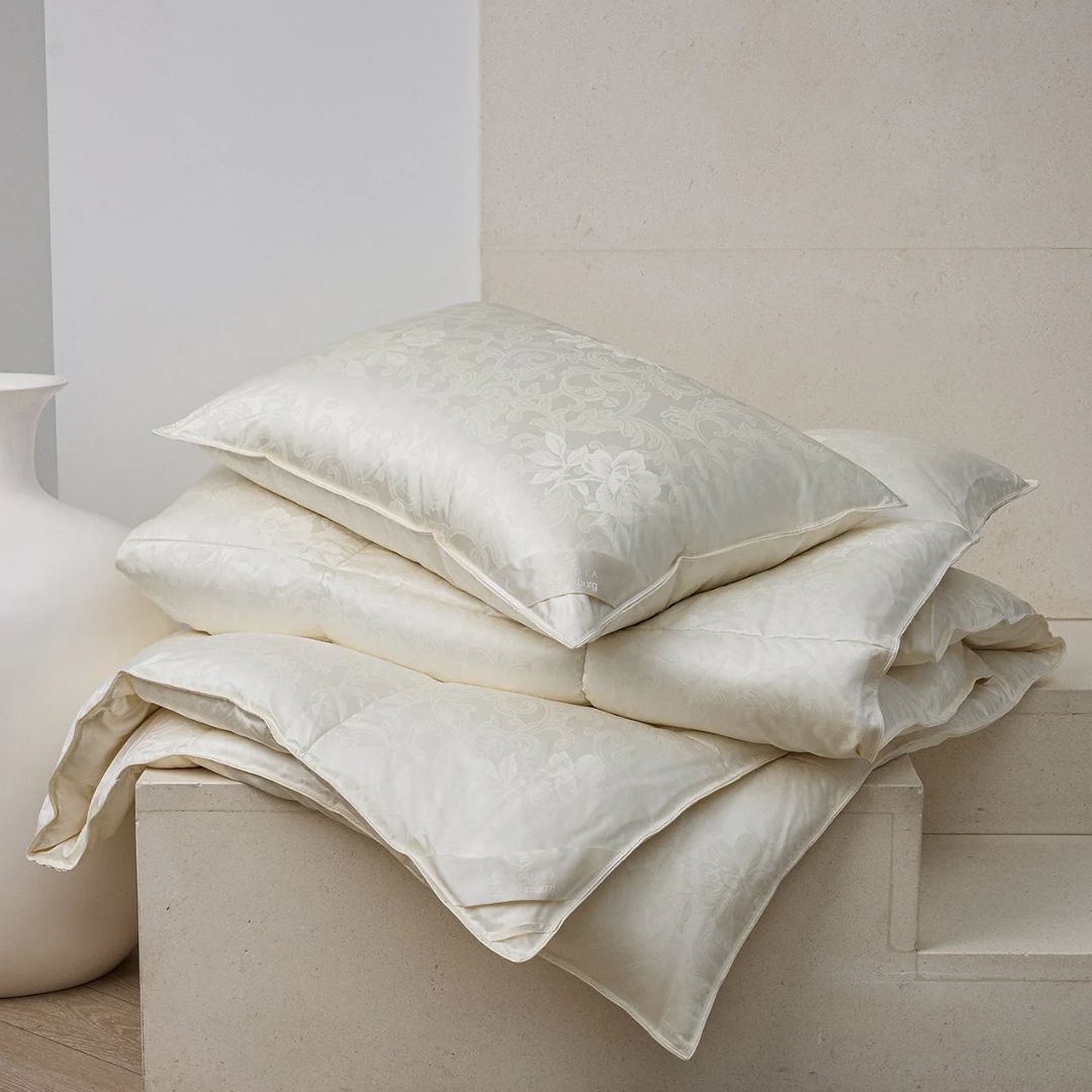 Three cream-colored, patterned silk comforters and a Scandia Home luxury down pillow stacked near a white vase.