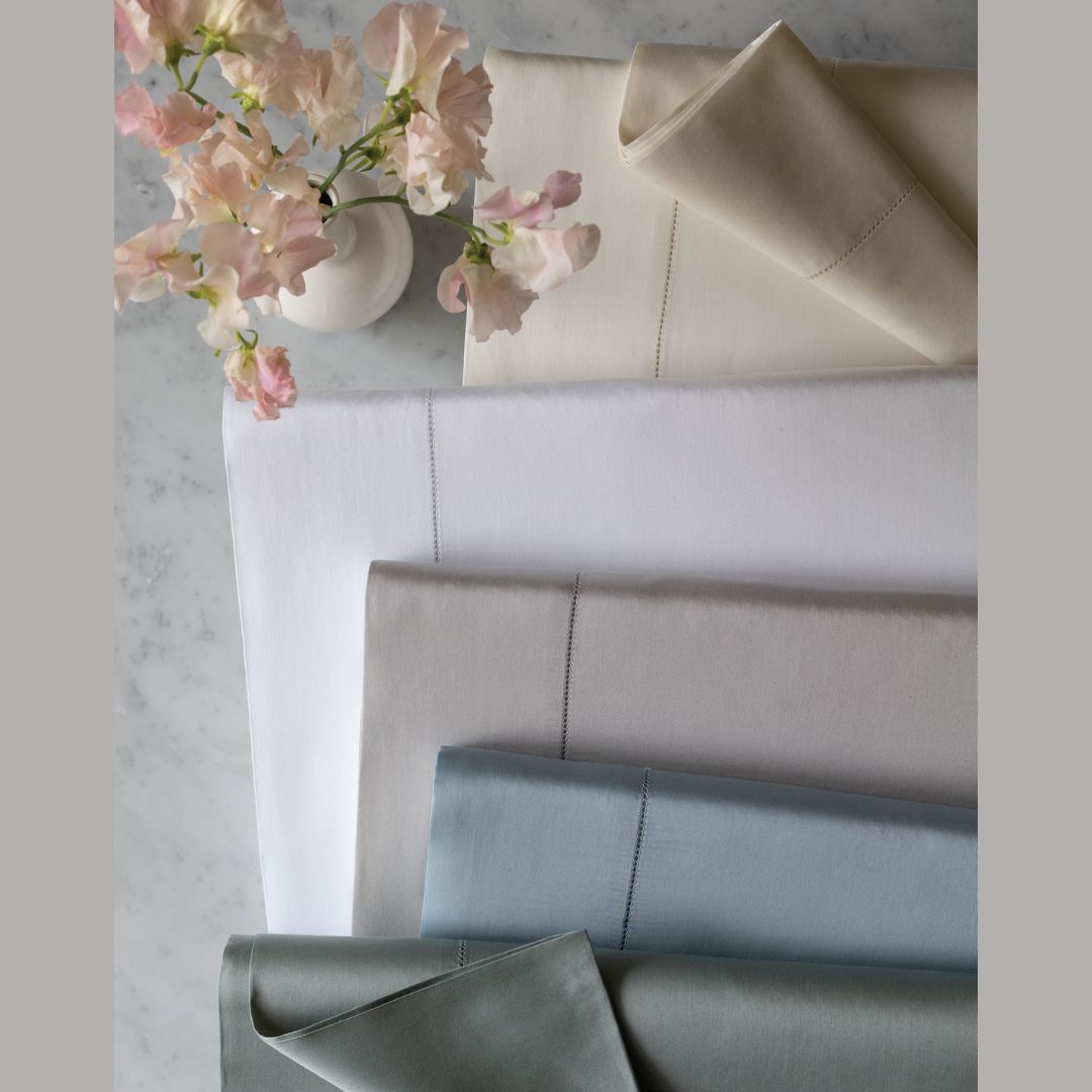 Five folded bedding sheets in neutral and cool tones arranged on a light marble surface beside a small white vase with pale pink flowers. The colors shown are White, Ivory, Heath, Rain, Sand, and Shadow.
