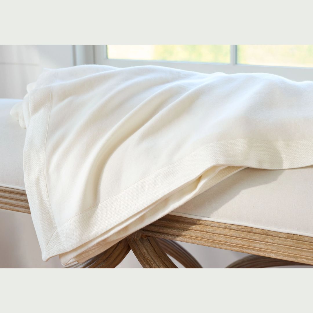 A folded Legna blanket is draped over a beige cushioned bench with wooden legs near a window.