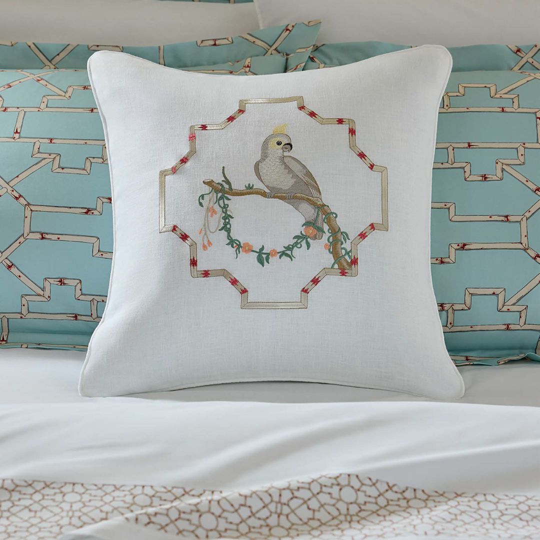 A decorative linen pillow with an embroidered bird perches on a bed with blue and patterned bedding.