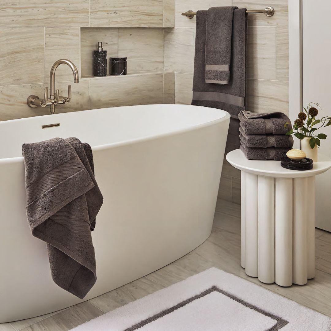 Modern bathroom with a white tub, gray Sferra Bello Nuovo Towels, small round table, and beige marble tile walls and floor.