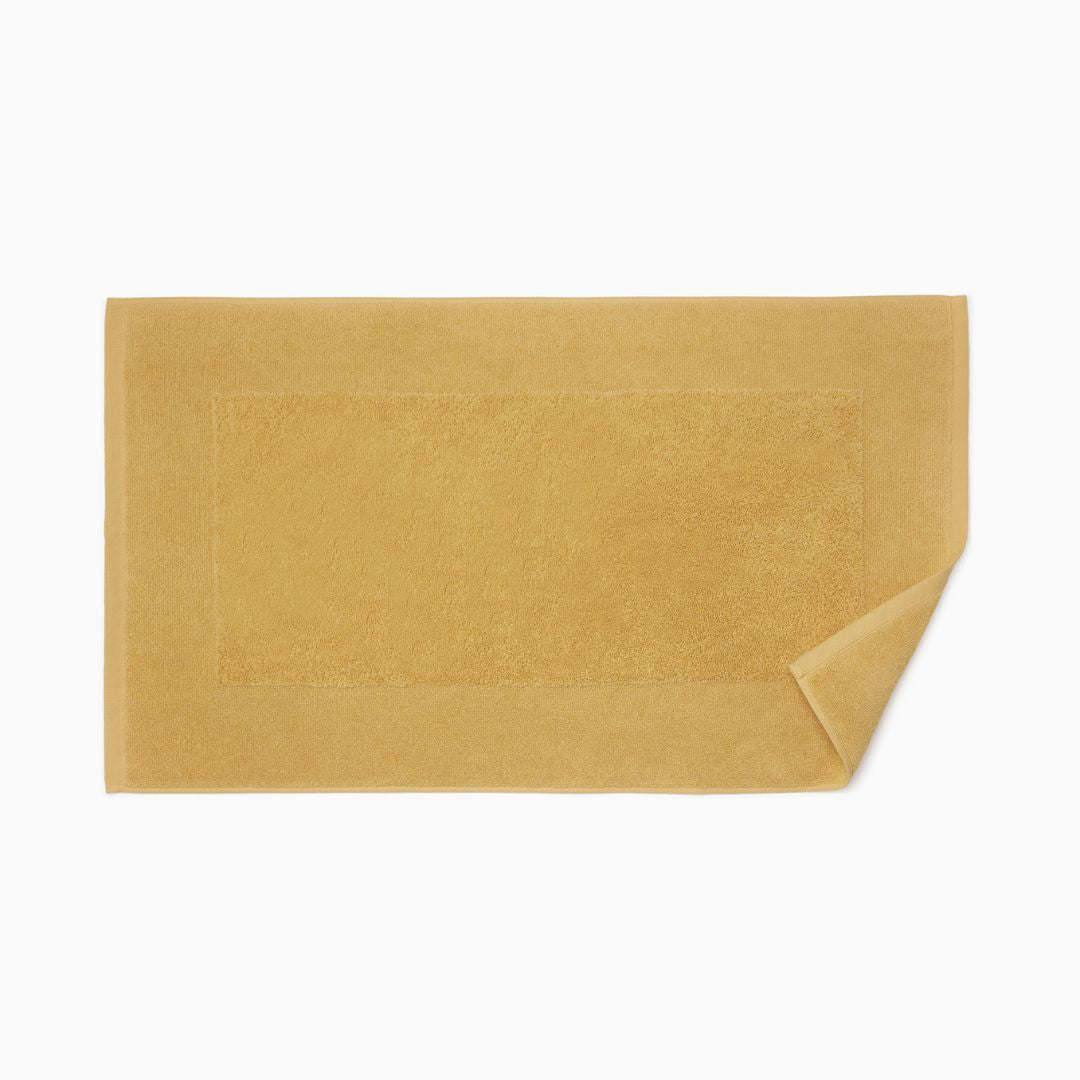 A rectangular mustard yellow bath mat, crafted from long-staple combed cotton, with one corner folded over.