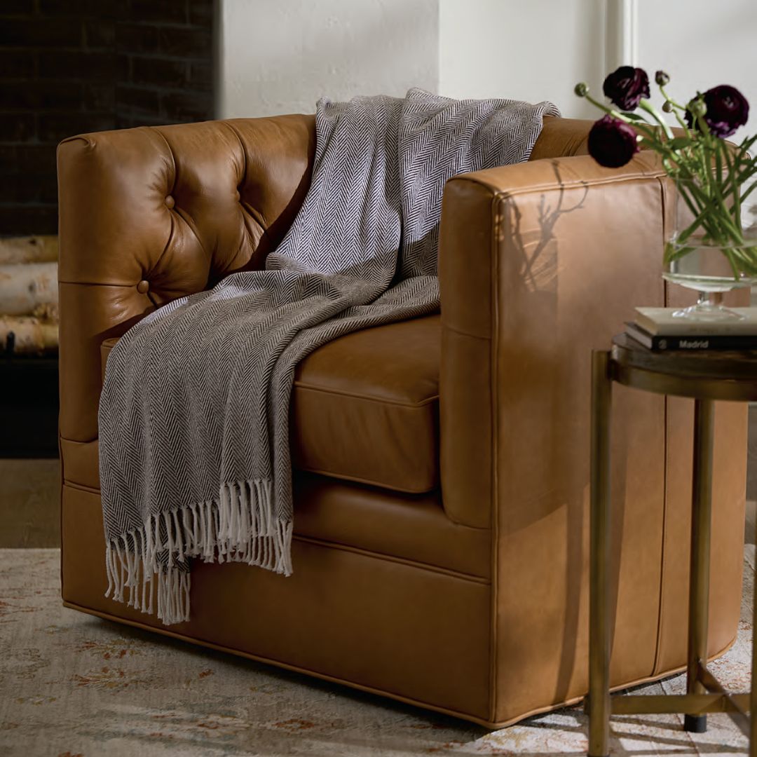 A brown leather armchair with a gray Celine Throw in herringbone weave, beside a round side table with flowers.