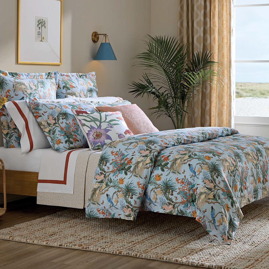A neatly made bed with Italian Riviera bedding, multiple pillows, a rug, a potted plant, and tan-curtained window.