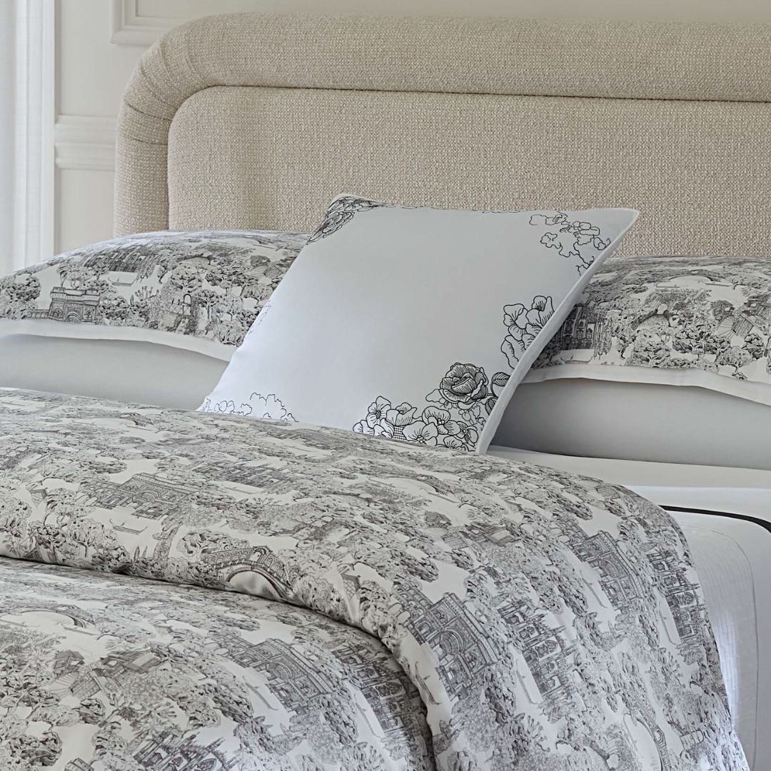 A bed with a light upholstered headboard and bedding in black and white designs, topped with a Sferra Fiorella Pillow.