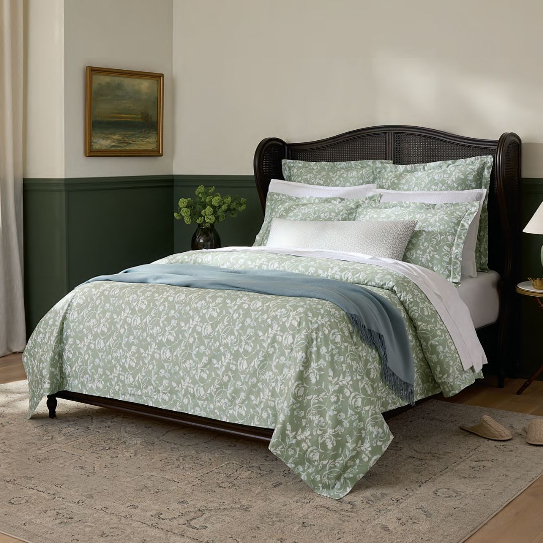 A neatly made bed with sage green bedding, a blue blanket, and a decorative pillow in a softly lit bedroom.