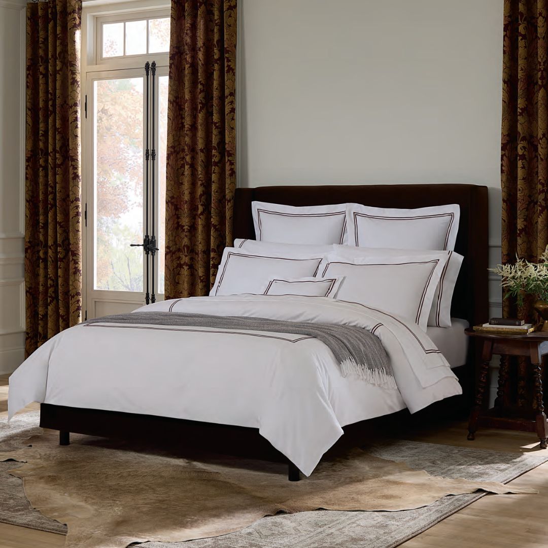 A neatly made bed with hotel bedding and brown trim, set in a bedroom with patterned curtains and a large window.