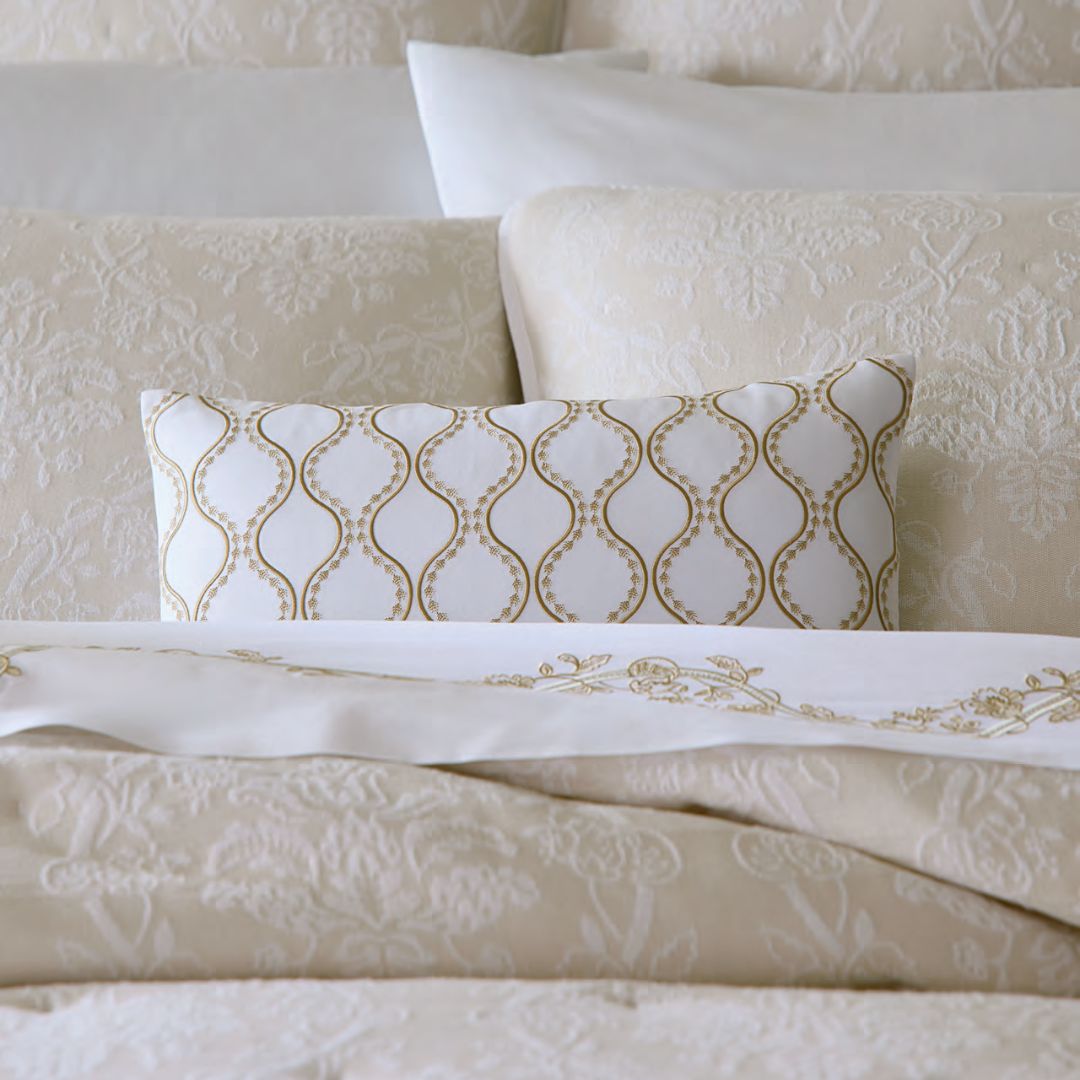 A bed with beige floral bedding and a Sferra x Scalamandre decorative pillow with a gold geometric design at the center.