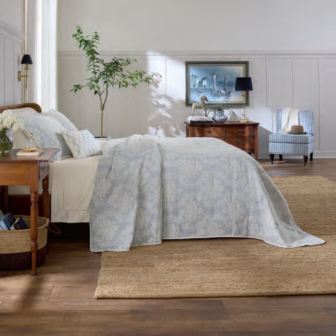 A neatly made bed with Sferra Isabella bedding, wooden furniture, a plant, and a painting in a bright bedroom.