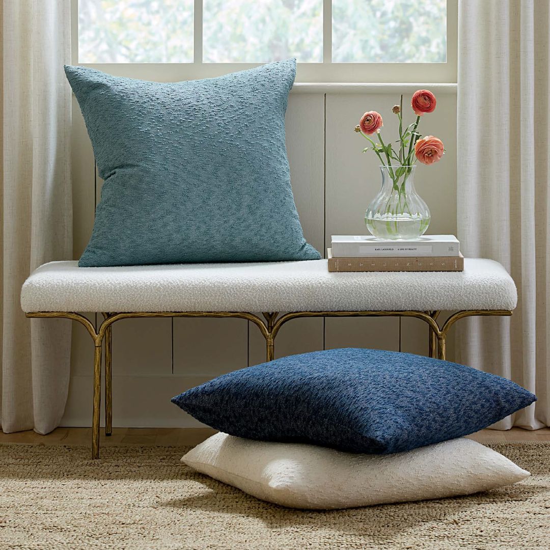 A blue jacquard texture pillow on a bench, a vase with flowers, books, and two pillows by the window.