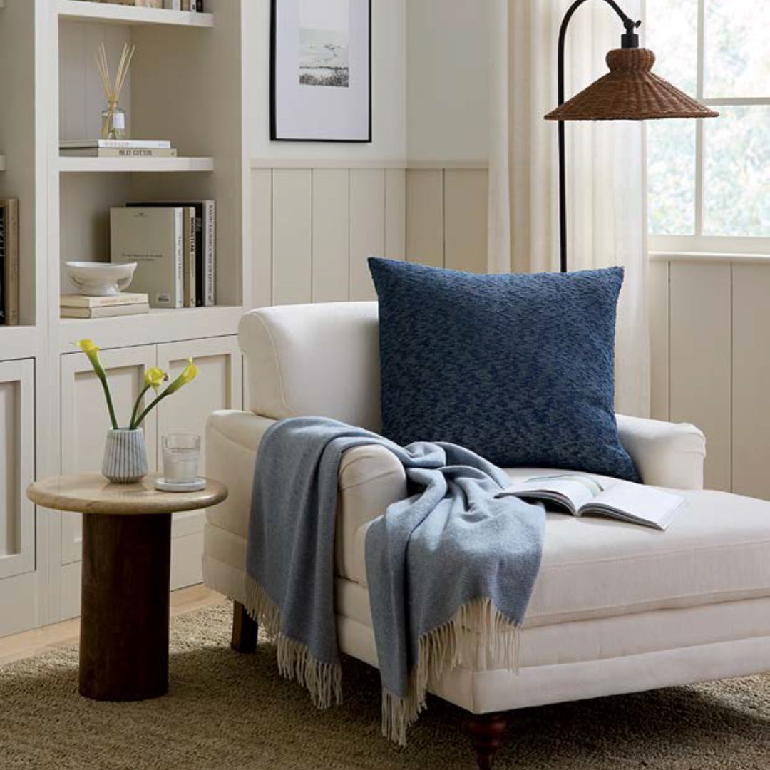 White armchair with a Sferra Lerici Decorative Pillow, blue throw, open book, and vase in a bright, cozy living room.