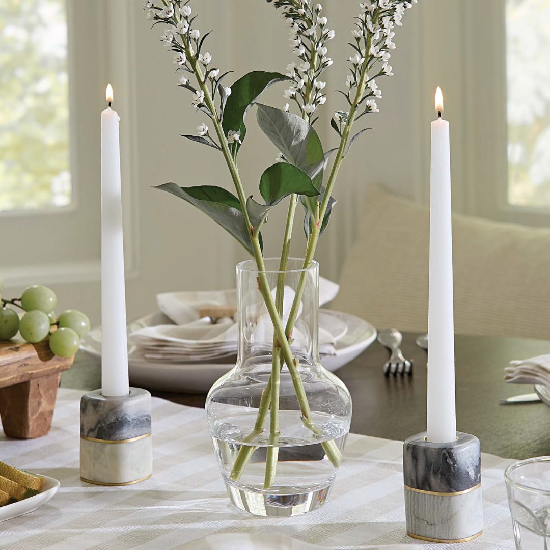A glass vase with leafy stems stands between two Sferra Lunario taper holders on a dining table set with plates and grapes.